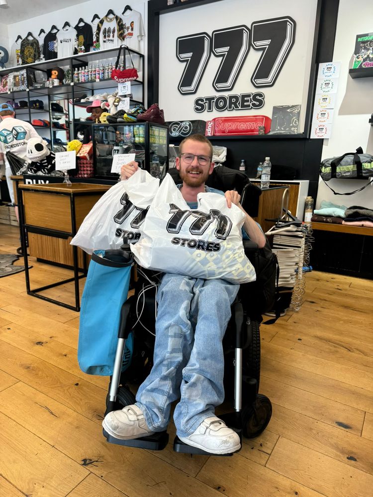 Tom in his powerchair in 777 Stores, grinning and holding up two big bags of shopping.