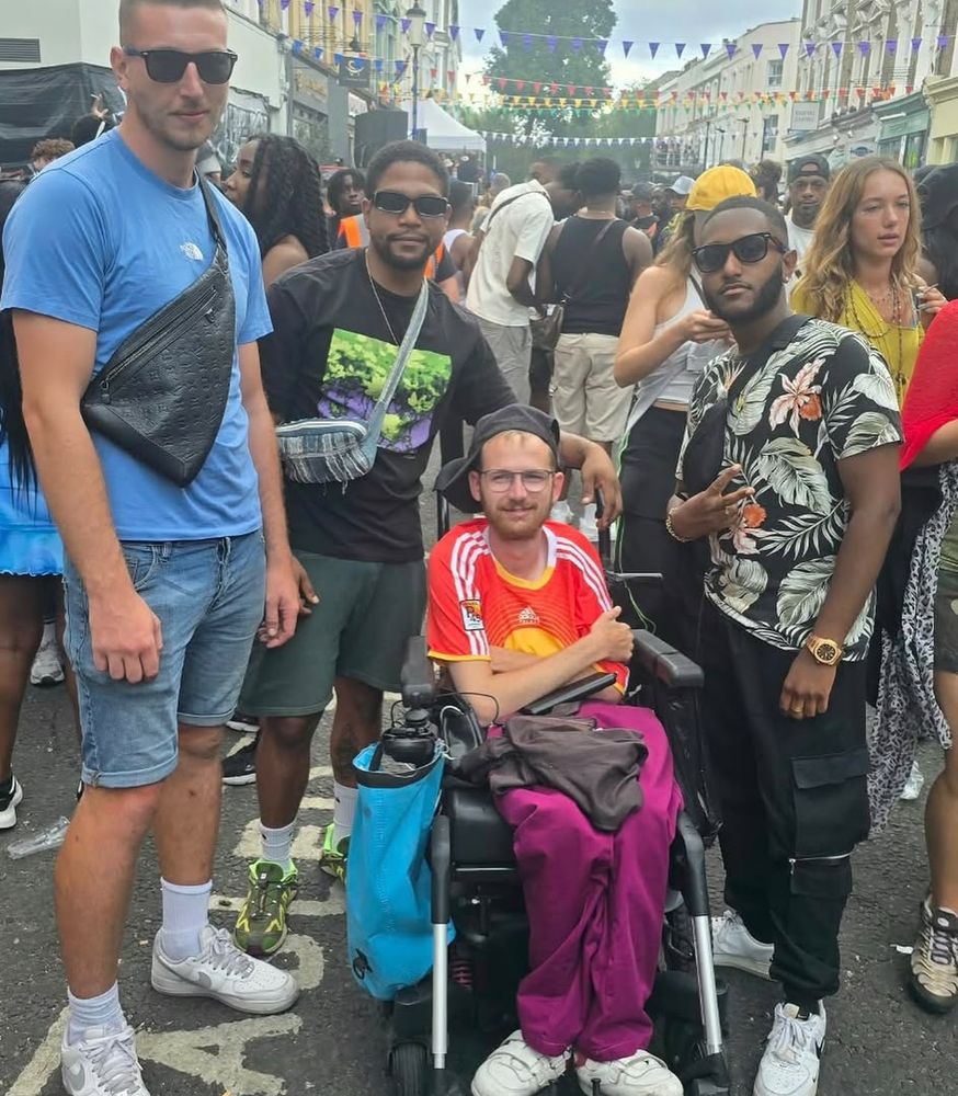 Tom at carnival in a red and yellow football shirt, sat in his powerchair in the street with three mates standing round him and bunting overhead.